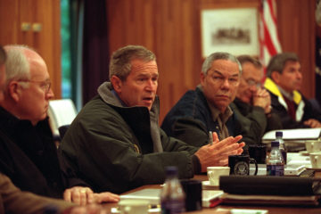 Photograph of George Bush sitting at a table with advisors including Dick Cheney and Colin Powell surrounding him.